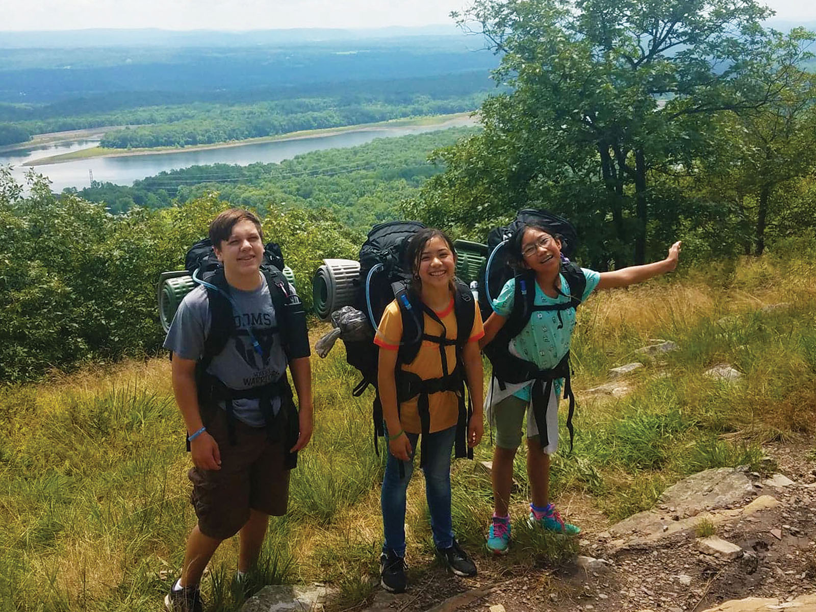 Three teen hikers with backpacks at the top of a rise overlooking a river