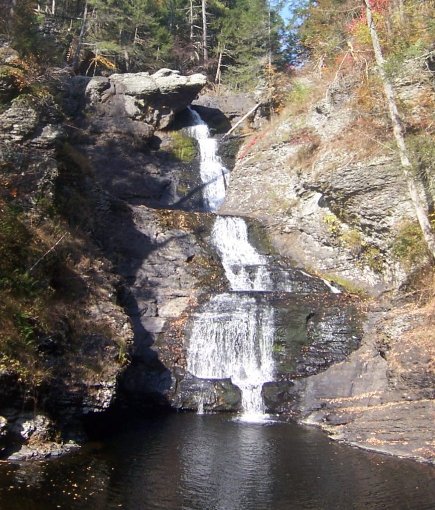 A tall waterfall over a rock face surrounded by autumn forest on both sides
