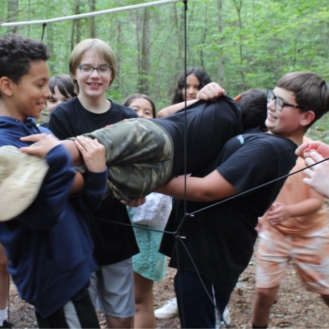 A group of boys passes one camper through a net designed to resemble a human-sized spiderweb in an activity on our Teambuilding Course.