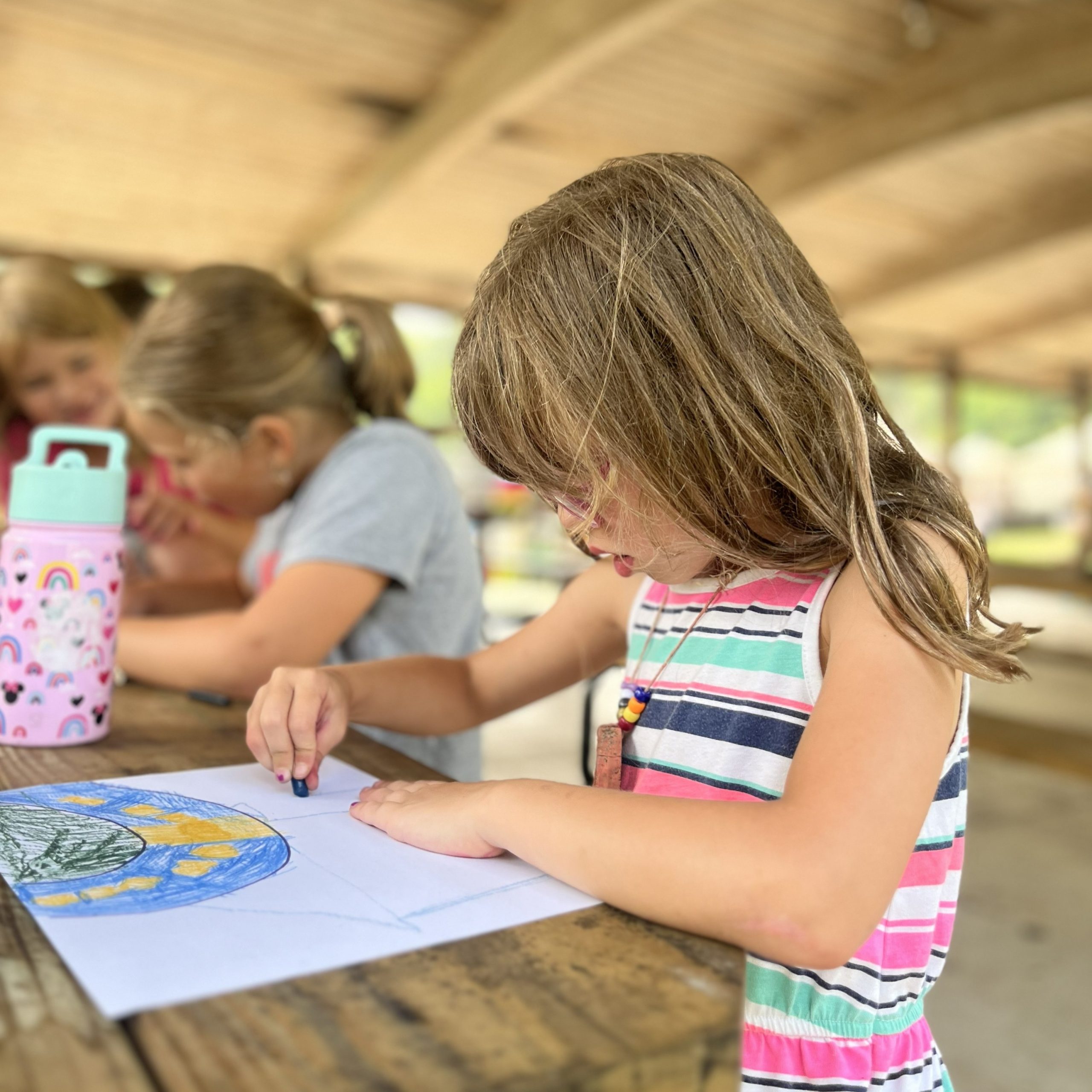 A young girl works with a crayon on paper to develop land alongside of a river as part of a summer camp activity.
