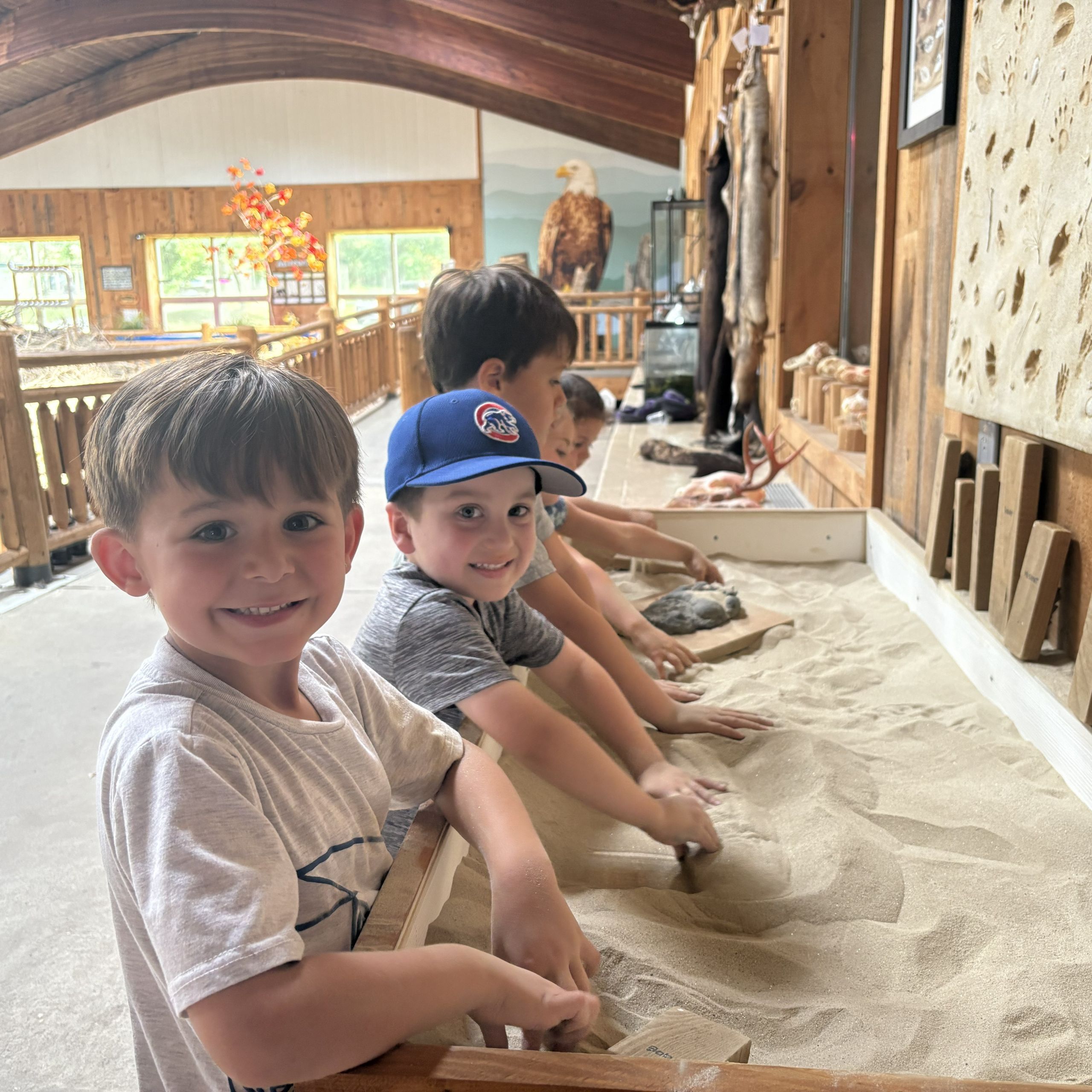A line of pre-school to kindergarten age campers investigate a sand pit with animal footprint track molds in PEEC's EcoZone. The two closest campers are smiling at the camera.