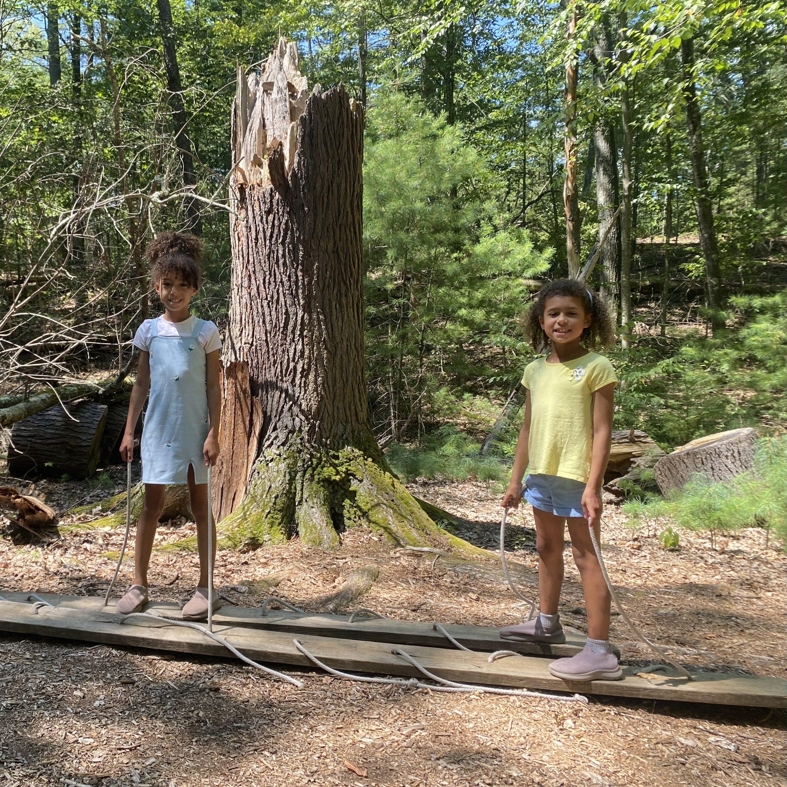 Two young girls smile at the camera as they stand on two wooden planks, holding attached ropes, as part of an activity on PEEC's teambuilding course.