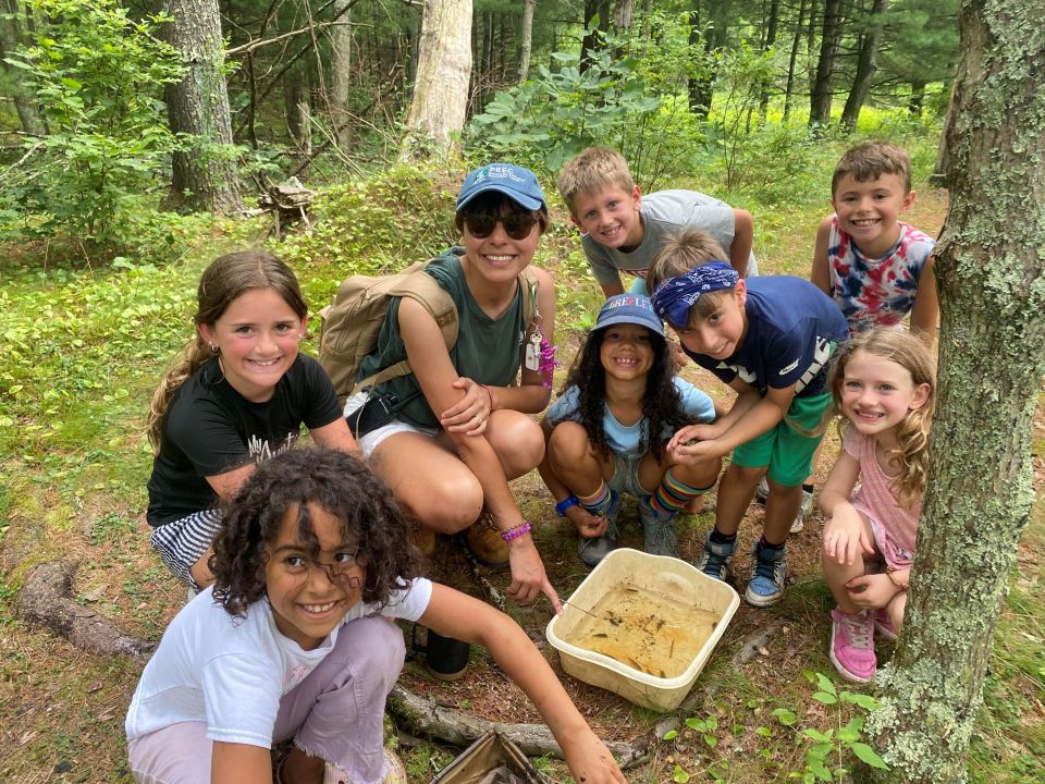 A group of elementary-aged campers and a PEEC instructor surround a bucket after collecting pond life. Everyone is smiling at the camera as they show off their findings.