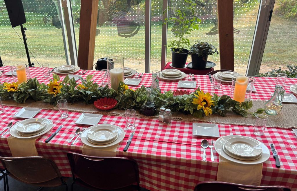 A long table with chairs set for dinner with red and white plaid picnic table tablecloths, a sunflower runner in the center, and tall candles amid the sunflowers